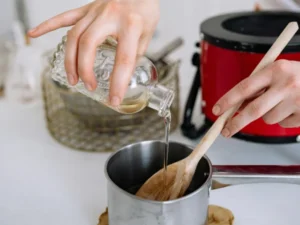 Image shows the hand of a woman pouring essential oil into a saucepan used for making wax melts. In the background is a red wax melter