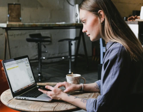 A woman carefully maps out her candle-making marketing strategy for an upcoming social media campaign. Seated at a small table, she works on her laptop, refining an Instagram template to showcase her scented candles and attract new followers. A strong cup of coffee sits beside her, fueling her creativity as she strategises her next move.