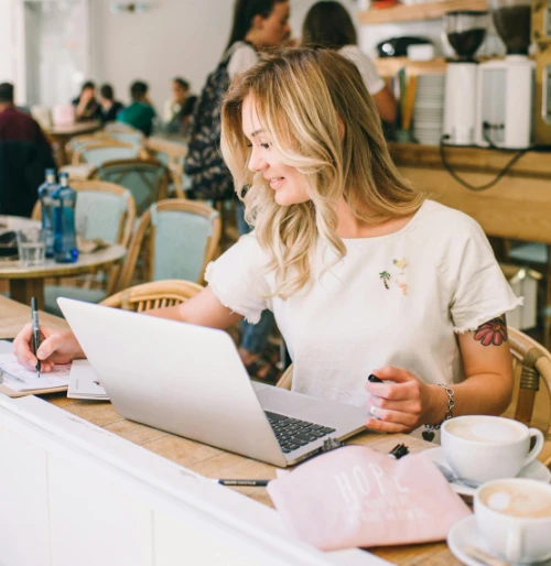 A woman smiles confidently as she carefully details her latest product designs into her candle making template in a busy café. Her laptop sits open beside her, aiding in her business launch preparations. Two cups of coffee rest on the table, while in the background, café patrons sit at tables and others place orders at the counter.