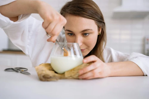 A skilled candle maker fastidiously trims the wooden wick of a white scented candle. She concentrates intently, to ensure she does not damage the wax or glass jar.